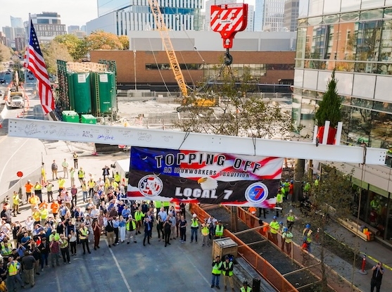a crane lifts a white beam with a pine tree, an American flag, and a banner for the Ironworkers Local 7 Union while a crowd of construction workers and other bystanders watches from the street