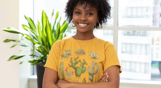 Chanell Mangum stands in a brightly lit room and smiles at the camera. She has her arms crossed and is wearing a yellow shirt with cacti on it.
