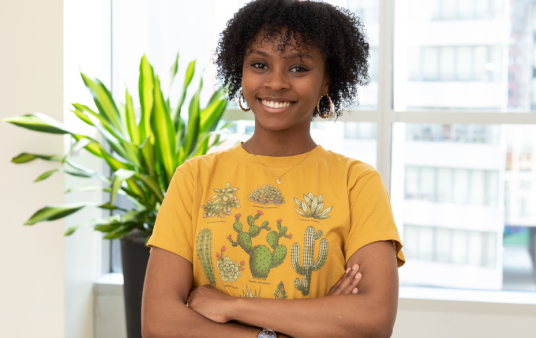 Chanell Mangum stands in a brightly lit room and smiles at the camera. She has her arms crossed and is wearing a yellow shirt with cacti on it.
